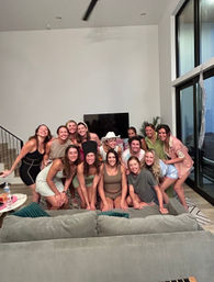 Cheerful group of women posing for a party group photo in a modern living room with large windows, a gray sectional sofa, and a TV.