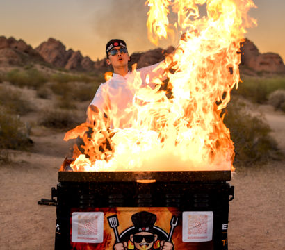 Hibachi chef performing a dramatic fire show at sunset over a portable grill in a rocky desert landscape, wearing sunglasses and a headband.