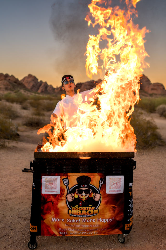 Hibachi chef performing a dramatic fire show at sunset over a portable grill in a rocky desert landscape, wearing sunglasses and a headband.