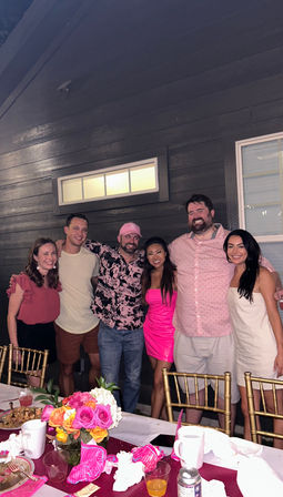 Group of six friends smiling and posing at an evening outdoor patio party against dark wood siding, with a table in the foreground featuring a bright pink and orange rose centerpiece, pink decorations, drinks and gold chairs.