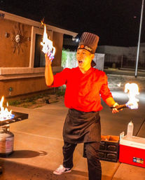 Hibachi chef in red jacket and tall hat performing a dramatic flaming cooking show on an outdoor patio at night, holding a torch and flaming spatula near a propane grill and cooler.