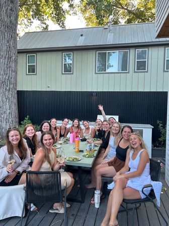 Group of friends laughing and toasting with wine around a long outdoor dining table on a wooden deck, salads and drinks on the table, with a large tree and modern two-story house in the background.