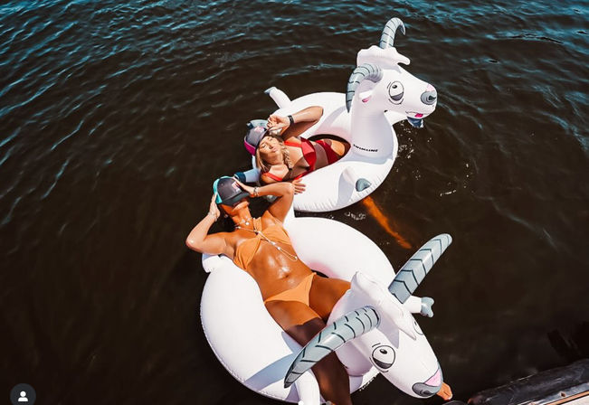 Two people lounging on white inflatable goat floats in dark water, wearing colorful bikinis and sun visors, relaxing and soaking up the sun