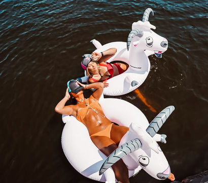 Two people lounging on white inflatable goat floats in dark water, wearing colorful bikinis and sun visors, relaxing and soaking up the sun