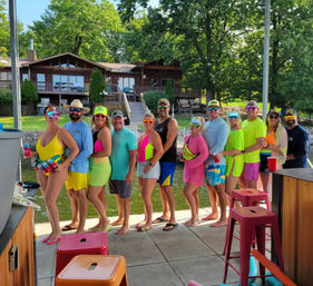 Group of adults in neon swimsuits and summer outfits lined up on a lakefront dock holding colorful drinks, with a wooden lakeside cabin and green trees in the background on a sunny summer day.