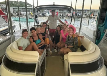 Eight people smiling on a cream-colored pontoon boat in a covered lake marina slip at sunset, casual summer outfits, a pink cowboy hat and an American flag visible