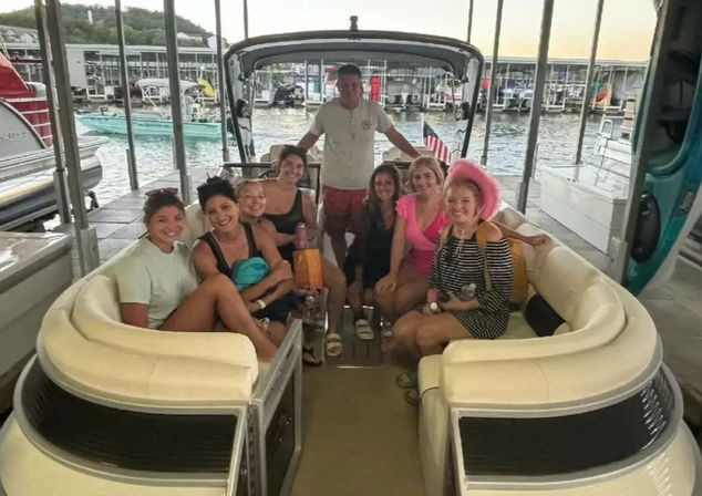 Eight people smiling on a cream-colored pontoon boat in a covered lake marina slip at sunset, casual summer outfits, a pink cowboy hat and an American flag visible