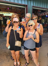 Five smiling women in sunglasses holding colorful frozen cocktails at an open-air summer bar with garage-style doors, wearing casual swimsuits and sandals.