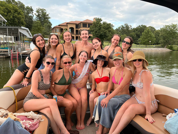Group of friends in swimsuits smiling on a pontoon boat at a lake with a large lakehouse, dock and tree-lined shore in the background — sunny summer boat party vibe.