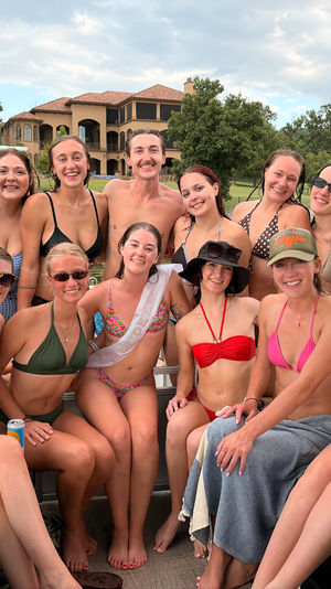 Group of friends in swimsuits smiling on a pontoon boat at a lake with a large lakehouse, dock and tree-lined shore in the background — sunny summer boat party vibe.