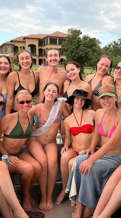 Group of friends in swimsuits smiling on a pontoon boat at a lake with a large lakehouse, dock and tree-lined shore in the background — sunny summer boat party vibe.