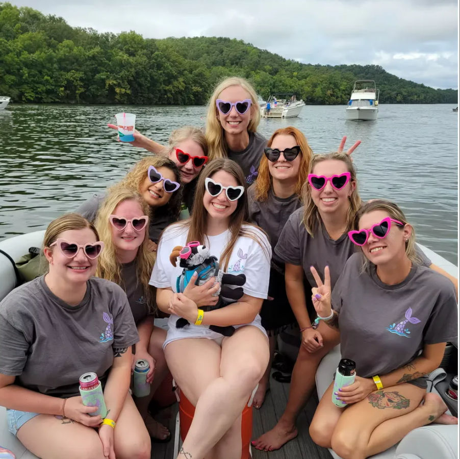 Group of women in colorful heart-shaped sunglasses on a pontoon boat enjoying a summer day on a tree-lined lake with other boats in the background