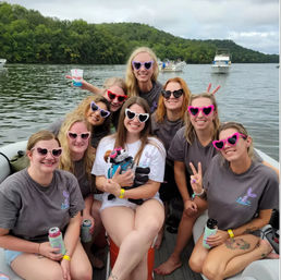 Group of women in colorful heart-shaped sunglasses on a pontoon boat enjoying a summer day on a tree-lined lake with other boats in the background