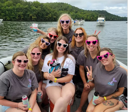 Group of women in colorful heart-shaped sunglasses on a pontoon boat enjoying a summer day on a tree-lined lake with other boats in the background