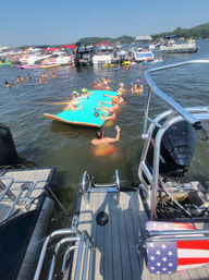 View from a boat deck at a busy lake party: people swimming and sipping drinks on a large turquoise floating mat surrounded by anchored pontoon and motorboats under a sunny summer sky.