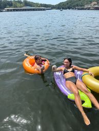 Two people relaxing on colorful inflatable tubes in a sunny lake near a marina, clinking canned drinks for a fun summer float day.