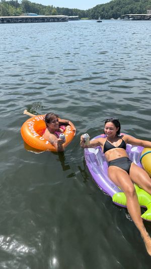 Two people relaxing on colorful inflatable tubes in a sunny lake near a marina, clinking canned drinks for a fun summer float day.