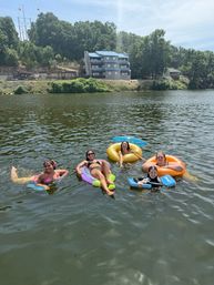 Five people smiling and relaxing on colorful inflatable tubes and a bodyboard in a sunny lake, with a tree-lined shoreline and a three-story blue-roofed building on the bluff in the background.