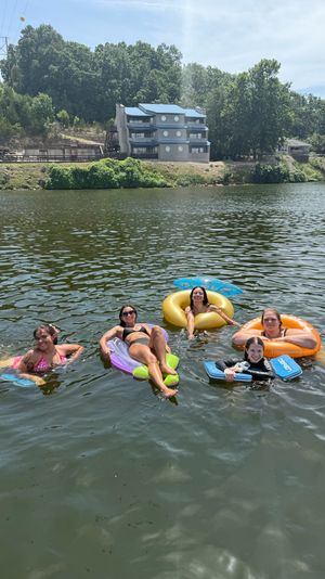Five people smiling and relaxing on colorful inflatable tubes and a bodyboard in a sunny lake, with a tree-lined shoreline and a three-story blue-roofed building on the bluff in the background.