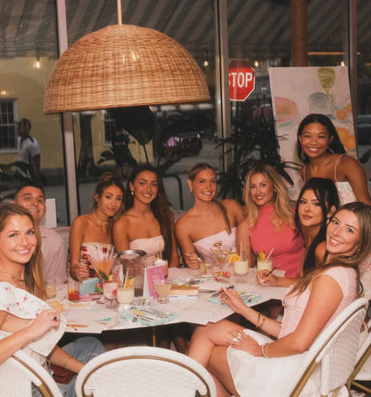 Smiling group of friends enjoying a girls’ night painting party at a cozy cafe — women around a table with cocktails, paint palettes and brushes under a large wicker pendant light, city street and stop sign visible through the window.