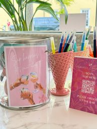 Sunlit sip-and-paint table with pink decor — metal tin showing a 'Sip happens' sign, two pink textured cups filled with colorful paint pens and a QR code menu card on a marble tabletop, plants and a car visible outside the window.