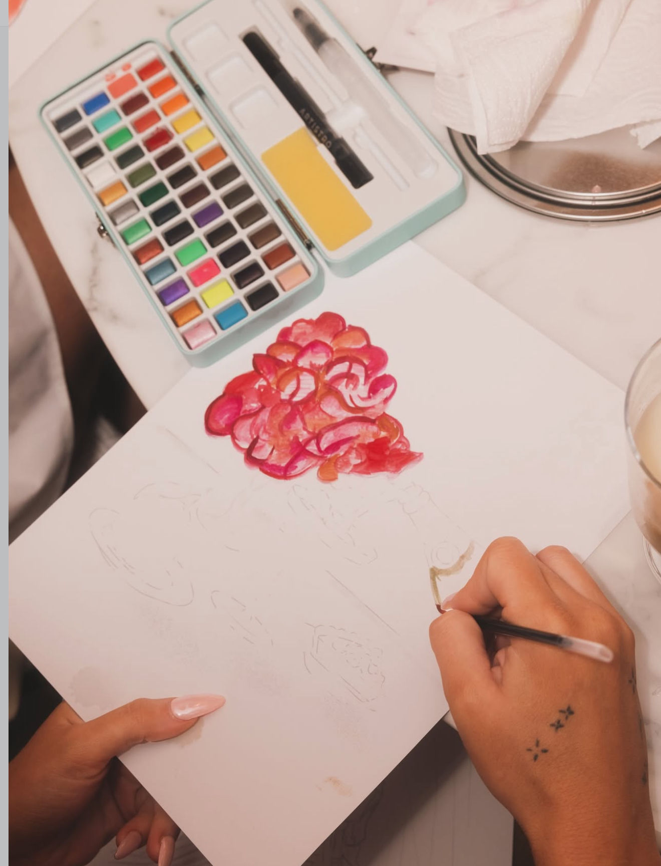 Top-down view of hands painting a pink and red watercolor floral cluster on paper, with a compact multicolor watercolor palette, brush, and a coffee cup on a marble tabletop.