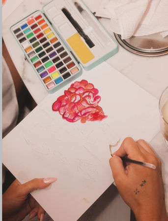 Top-down view of hands painting a pink and red watercolor floral cluster on paper, with a compact multicolor watercolor palette, brush, and a coffee cup on a marble tabletop.