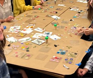 Group craft workshop around a kraft-paper-covered table with pressed flowers, floral stickers, glue, toothpick jars and hands arranging DIY botanical art