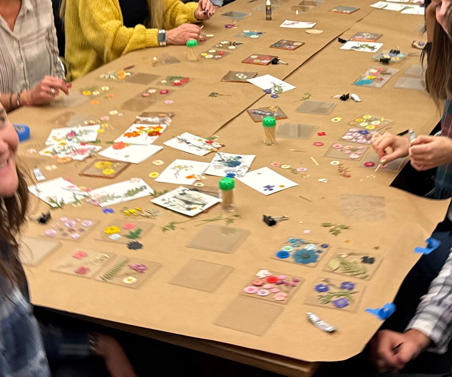 Group craft workshop around a kraft-paper-covered table with pressed flowers, floral stickers, glue, toothpick jars and hands arranging DIY botanical art