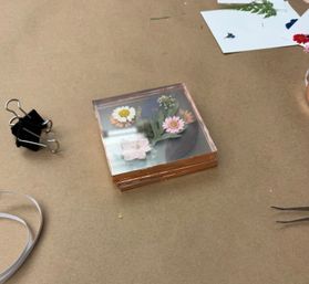 Stack of square clear resin coasters with colorful pressed daisies and leaves on a brown kraft paper craft table, surrounded by a binder clip, ribbon and tweezers.