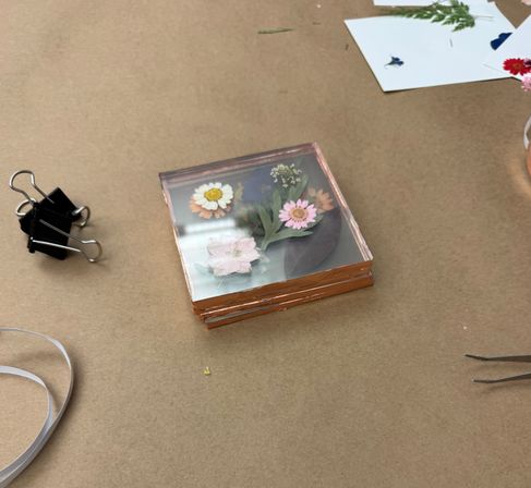 Stack of square clear resin coasters with colorful pressed daisies and leaves on a brown kraft paper craft table, surrounded by a binder clip, ribbon and tweezers.