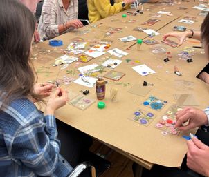 Busy DIY craft table with people arranging pressed flowers into clear acrylic coaster squares on kraft paper, with tweezers, tacky glue, toothpicks and binder clips nearby.