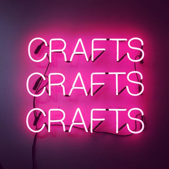 Triple pink neon 'CRAFTS' sign mounted on a wall, bold magenta glow and retro craft-studio decor.