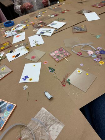 Tabletop view of a DIY pressed-flower craft workshop: assorted dried flowers, clear resin tiles and coasters, white cards, ribbon and glue scattered on brown craft paper.