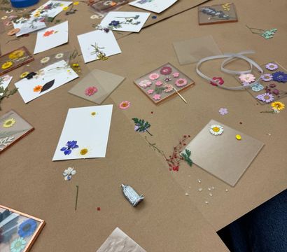 Tabletop view of a DIY pressed-flower craft workshop: assorted dried flowers, clear resin tiles and coasters, white cards, ribbon and glue scattered on brown craft paper.
