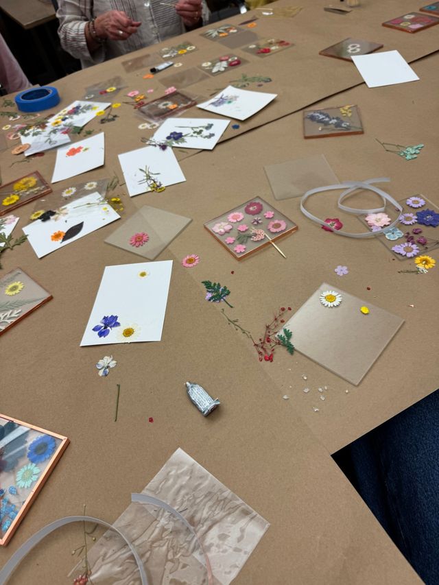 Tabletop view of a DIY pressed-flower craft workshop: assorted dried flowers, clear resin tiles and coasters, white cards, ribbon and glue scattered on brown craft paper.