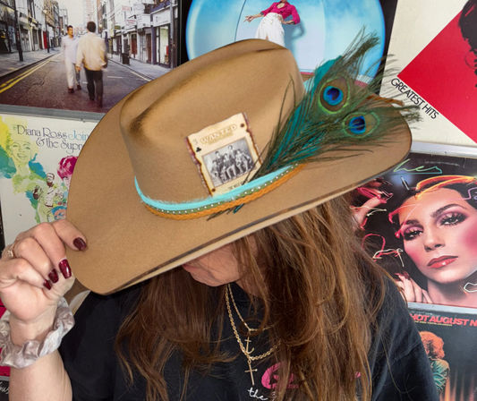 Close-up of a person tipping a tan cowboy hat decorated with peacock feathers, turquoise trim and a vintage 'wanted' card, set against a colorful wall of retro vinyl album covers.