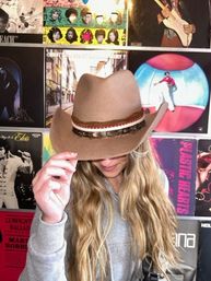 Person with long blonde hair playfully tipping a brown cowboy hat in front of a colorful wall of vinyl album covers, retro music-store vibe.