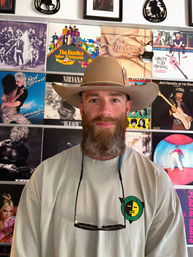 Bearded man wearing a tan cowboy hat with sunglasses hanging from his neck posing in front of a colorful wall of vinyl album covers in a retro record-shop style display.