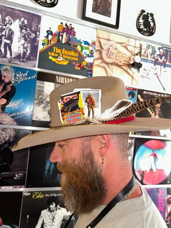 Profile of a bearded man in a tan wide-brim hat with feather and vintage card pin, standing in front of a colorful vinyl album wall in a record shop