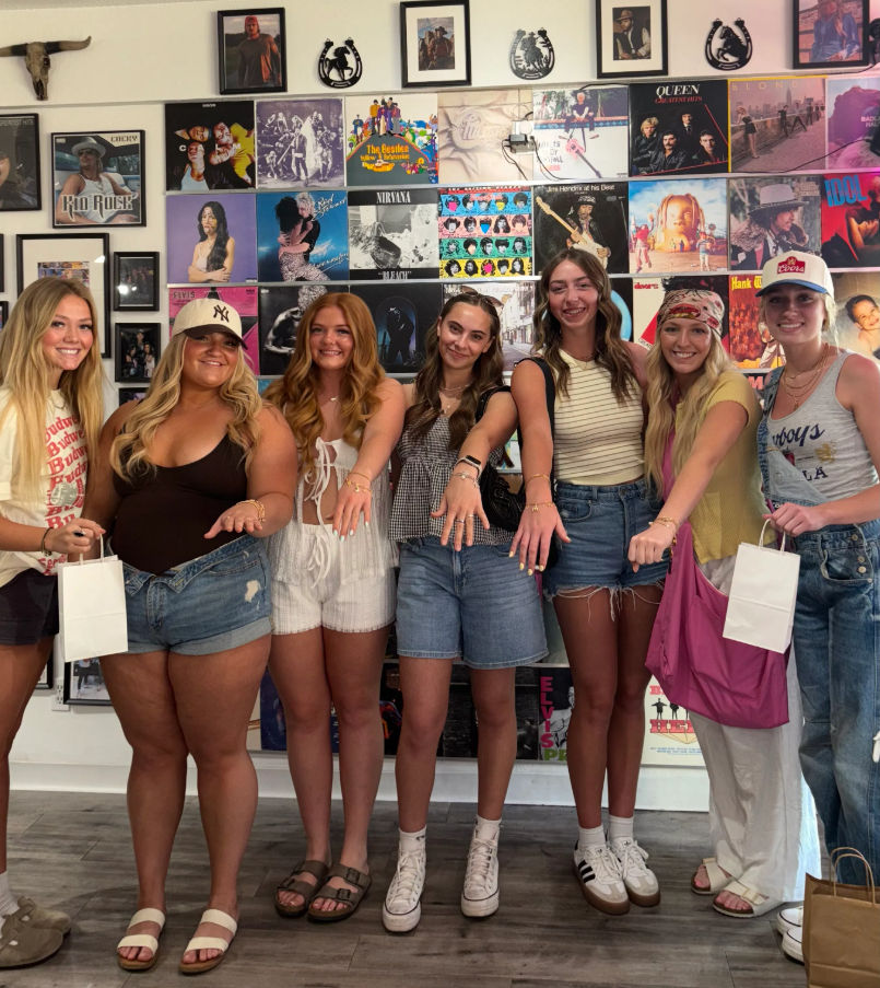 Seven young women smiling and posing inside a retro record shop with a colorful wall of vinyl album covers, showing off rings and summer casual outfits.
