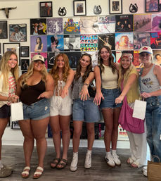 Seven young women smiling and posing inside a retro record shop with a colorful wall of vinyl album covers, showing off rings and summer casual outfits.