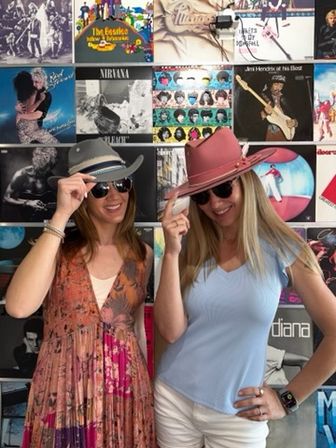 Two smiling women in sunglasses tipping a pink and a gray hat while posing in front of a colorful vinyl record wall backdrop — playful music-themed photo.