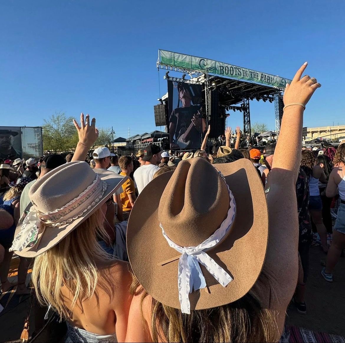 Outdoor country music festival crowd with cowboy hats and raised hands facing a main stage and large video screen under a clear blue sky