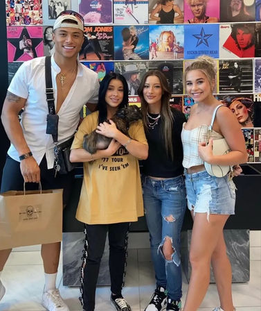 Four friends smiling and posing in front of a colorful vinyl album wall at a music shop, one cradling a small brown puppy; casual summer outfits with denim shorts, ripped jeans and an oversized tee, plus a shopping bag visible.