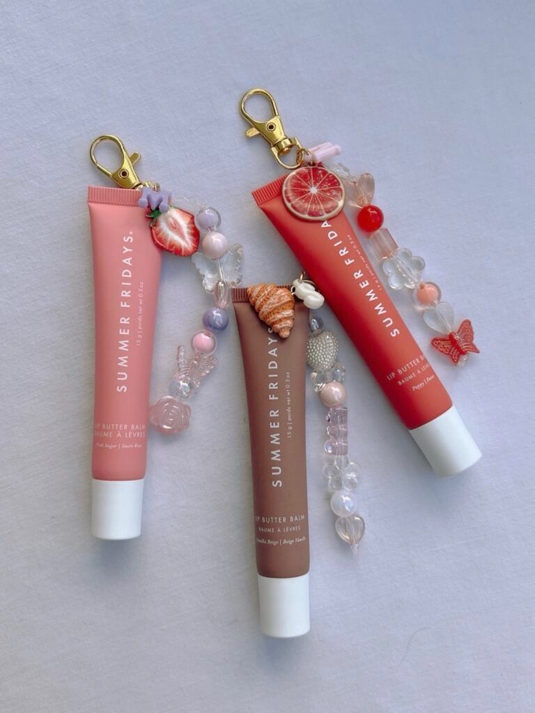 Flat lay of three colorful lip balm tubes with gold clasp keychain charms — pink, coral and nude tubes decorated with strawberry, citrus slice, seashell and pastel beads on a pale fabric background