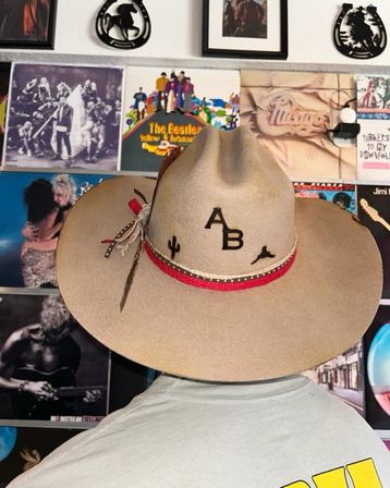 Back view of person wearing a beige western cowboy hat with "AB" monogram, cactus and longhorn icons, red braided band and feather, in front of a wall of colorful vintage record album covers.