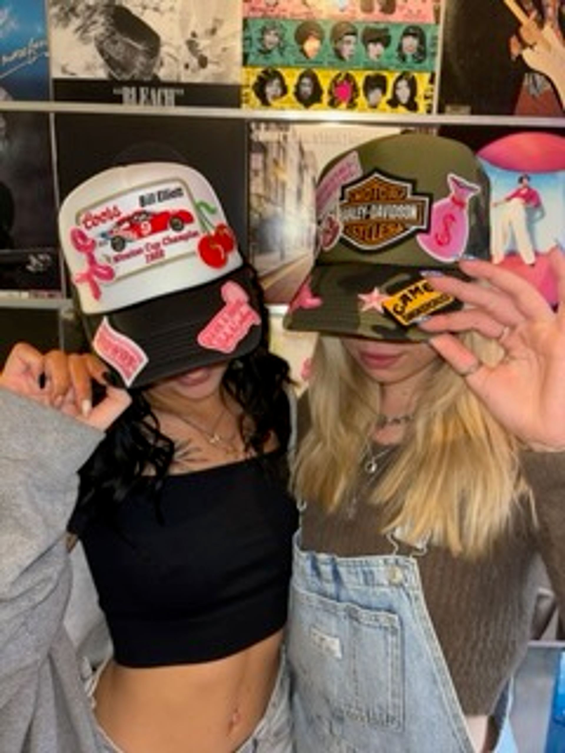 Two people posing in front of a colorful vinyl record wall, each wearing a decorated trucker hat—one in a black crop top and the other in denim overalls—fun, retro music-themed scene.