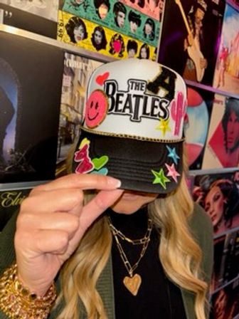 Person tipping a colorful The Beatles trucker hat with pink smiley, cactus and star stickers; long blonde waves, layered gold necklaces with heart pendant, retro vinyl album cover wall backdrop.