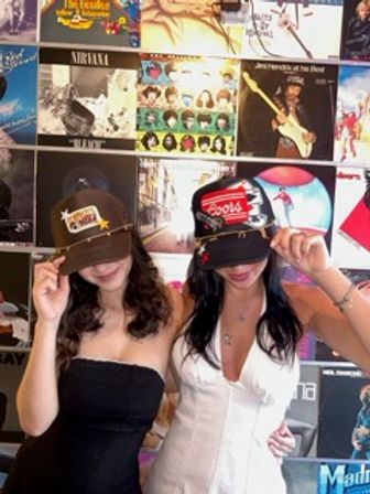 Two women wearing playful baseball caps pose side-by-side in front of a colorful wall of vinyl album covers at a record shop, tilting their hats down and smiling.